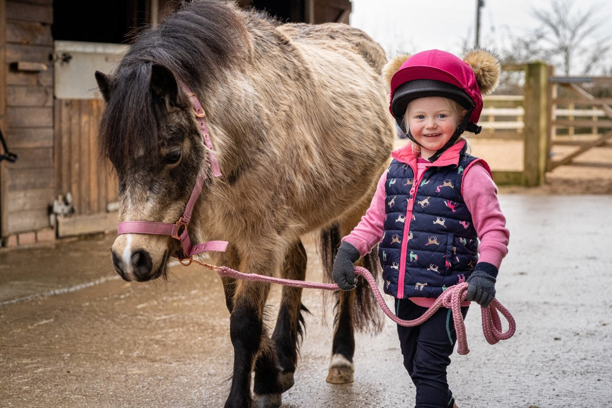 Smiling young rider leading a shaggy pony beside a stable